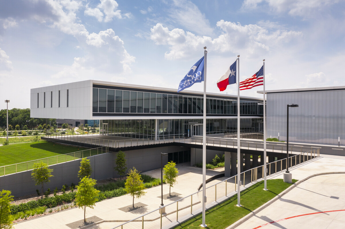 Photo of Collin College's Technical Campus located in Allen, Texas. The building is a multistory structure with a cantilevered third floor overhanging a dining area. A driveway to an underground garage is visible, as are greenspaces, and flags for the nation, the state, and Collin College.