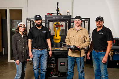 Welding student Sage Jeffery, Professor Cesar Lopez Maldonado, welding student Wyatt Jordan, and Professor Landon LaRocque stand in front of Collin College’s current automated welder. A grant will allow for the purchase of additional automated welders for student use and curriculum development.